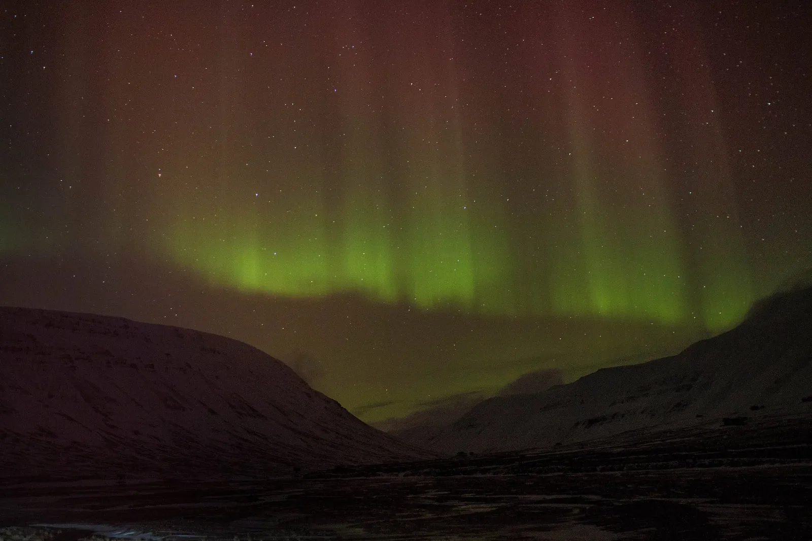Curtains of green and red northern lights rising above dark, snow-covered mountains under a starry sky