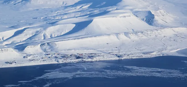 Barentsburg seen from a distance, a thin line of buildings along the shore of a dark fjord beneath broad snow-covered slopes
