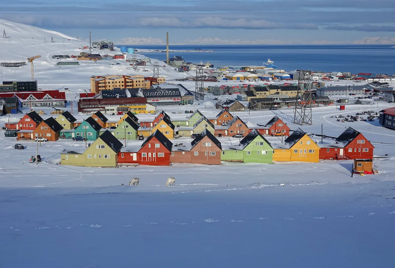 A row of brightly painted wooden houses in red, yellow, green and blue on the snow in Longyearbyen, with the fjord behind