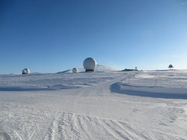 White radar domes of the Svalbard Satellite Station scattered across a snow-covered mountain plateau under a clear blue sky