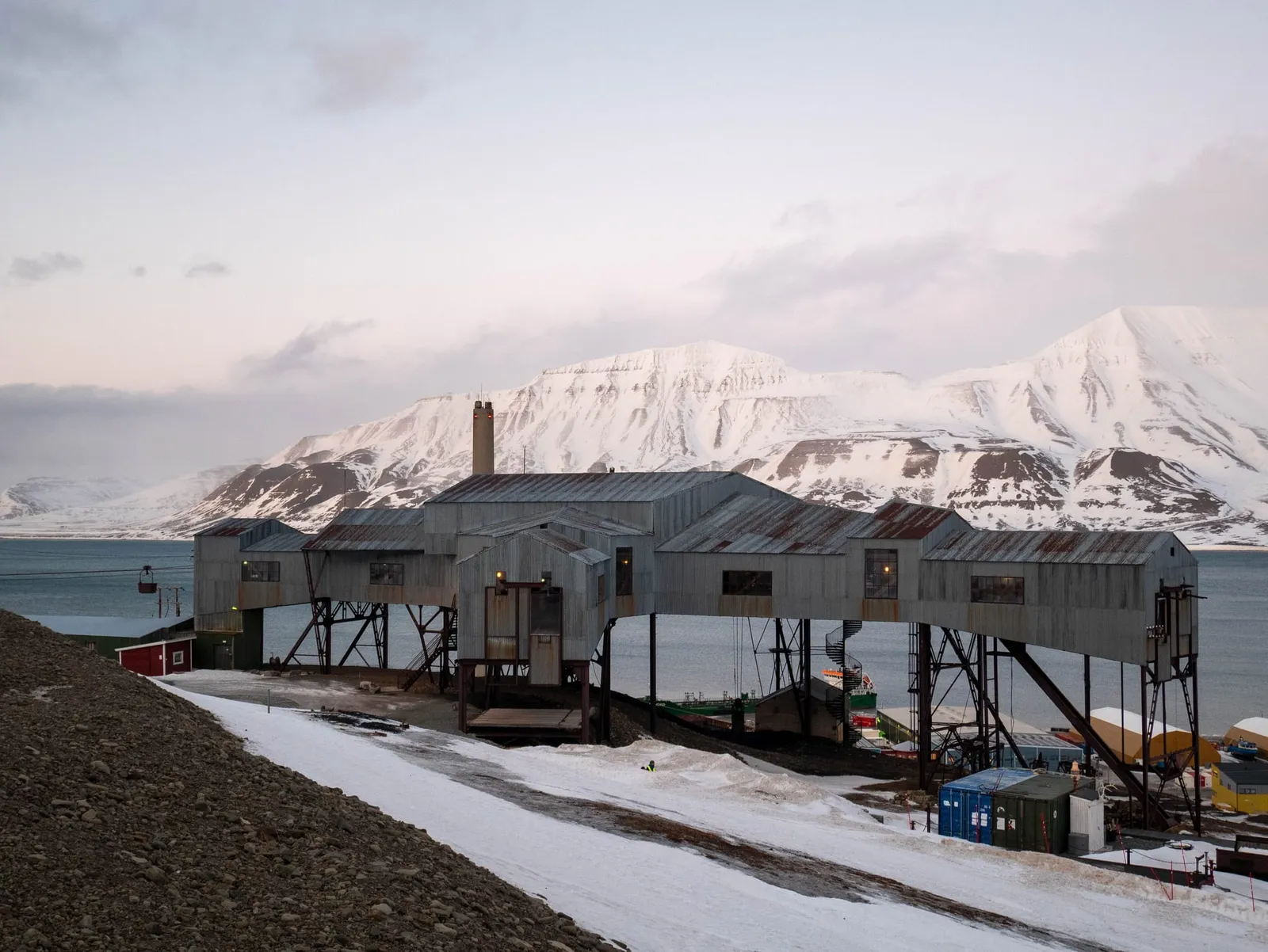 The grey timber buildings of the old coal cableway centre standing on stilts above the town, with snow-covered mountains across the fjord behind