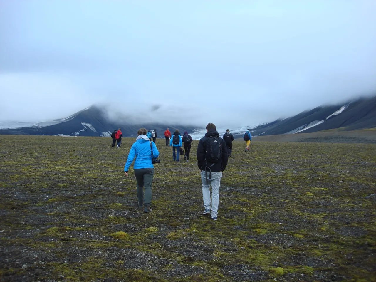 A line of hikers in rain jackets walking across a flat, mossy mountain top towards low cloud