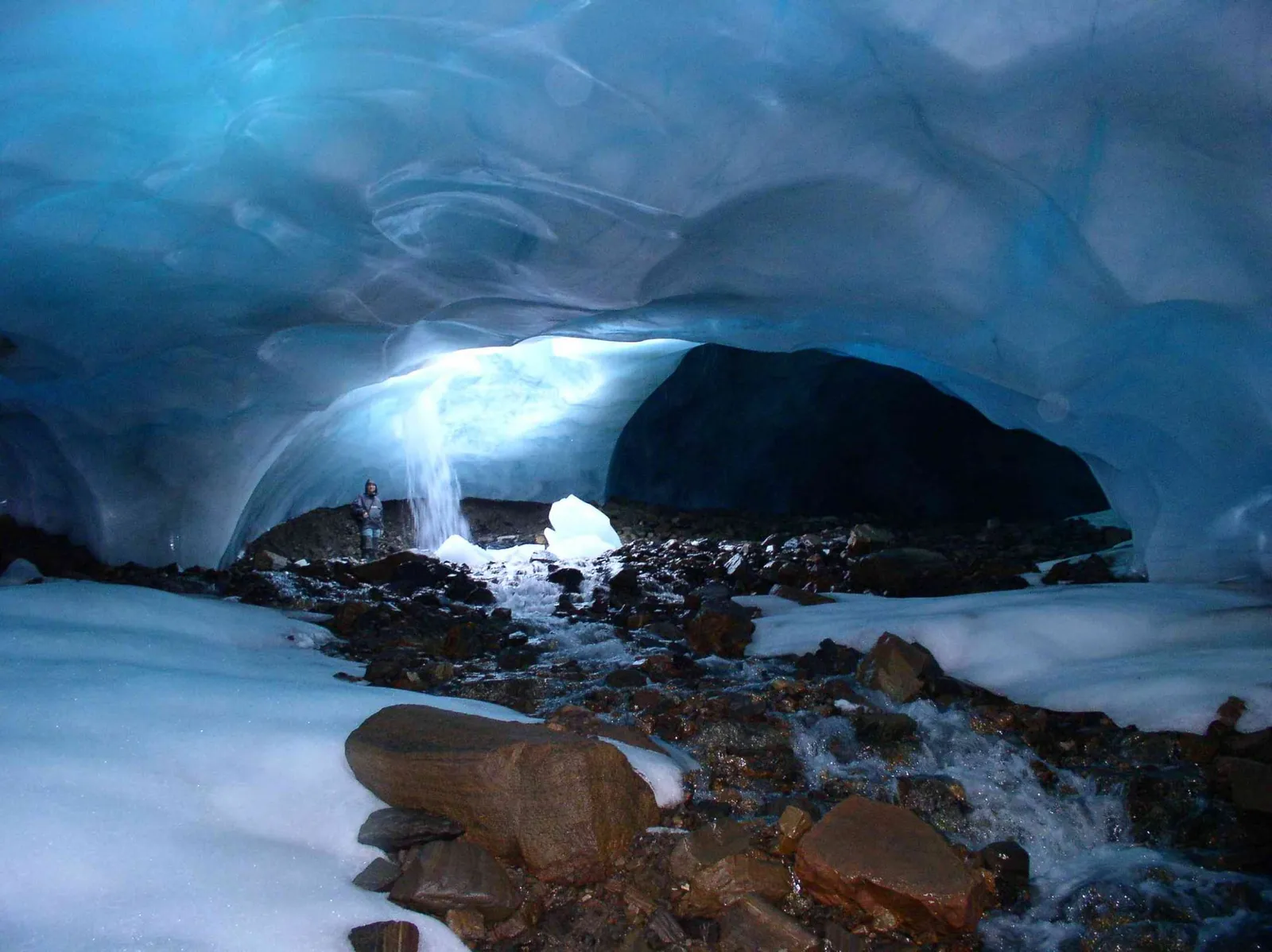 Inside a glacier ice cave, with a blue, rippled ice ceiling arching over a meltwater stream running between rocks
