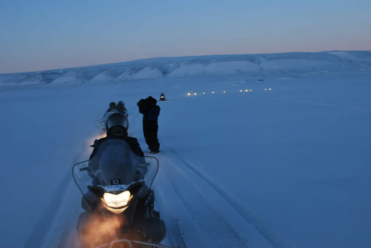 Snowmobiles with their headlights on crossing snow-covered tundra in deep blue twilight