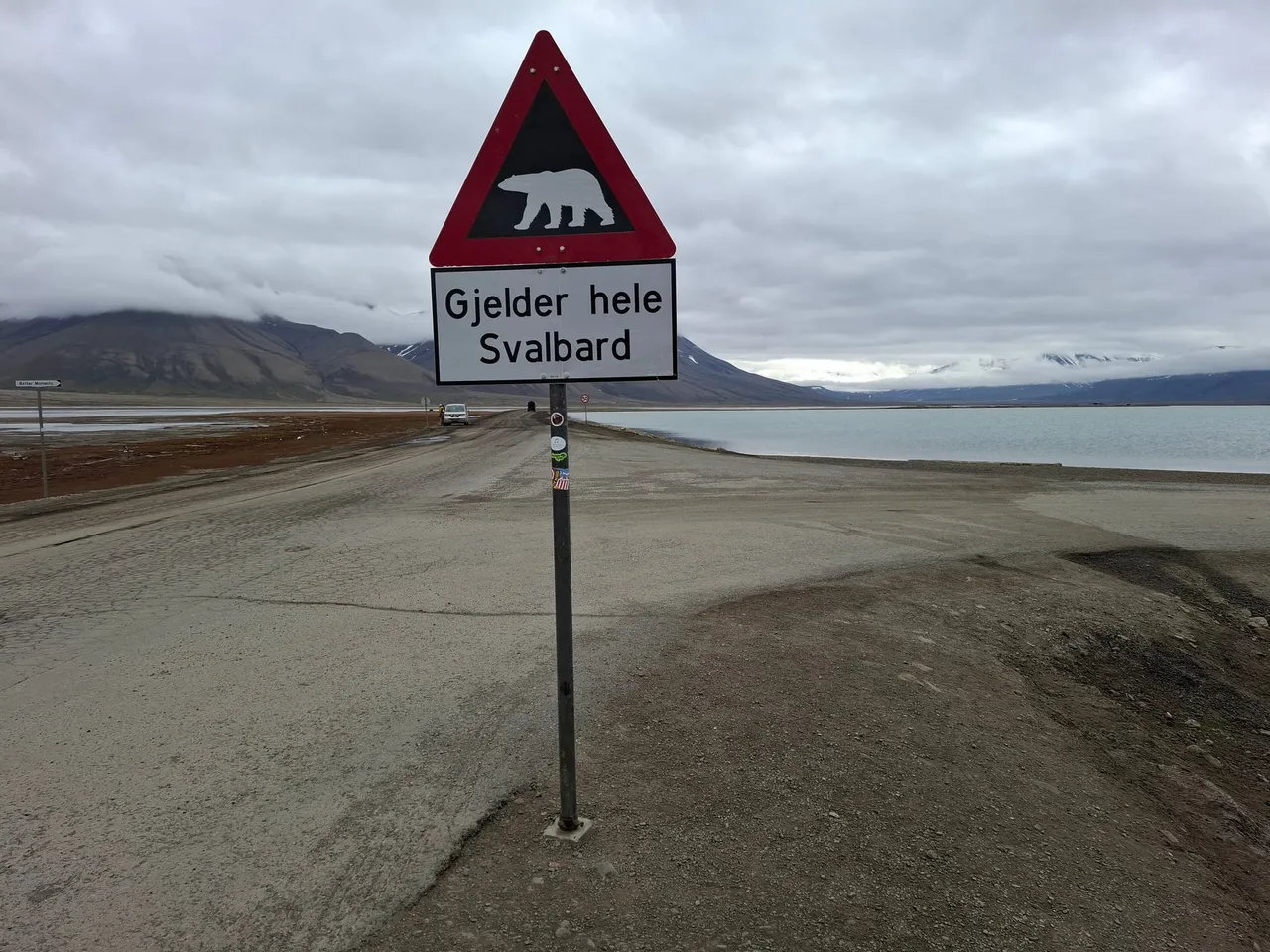 A red-bordered triangular road sign showing a polar bear, with the words Gjelder hele Svalbard, beside a gravel road along the fjord