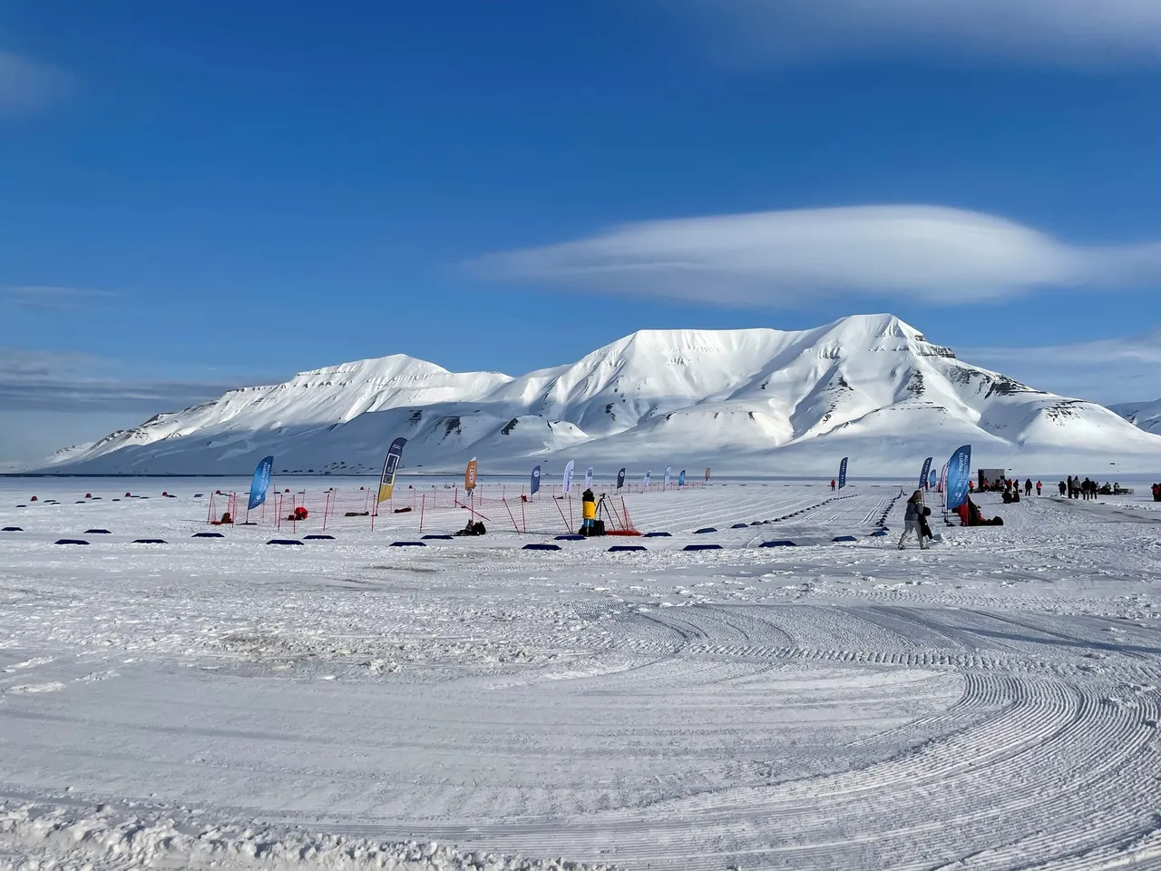 A wide, groomed snow arena with flags and tents in front of snow-covered mountains under a blue sky with lenticular clouds
