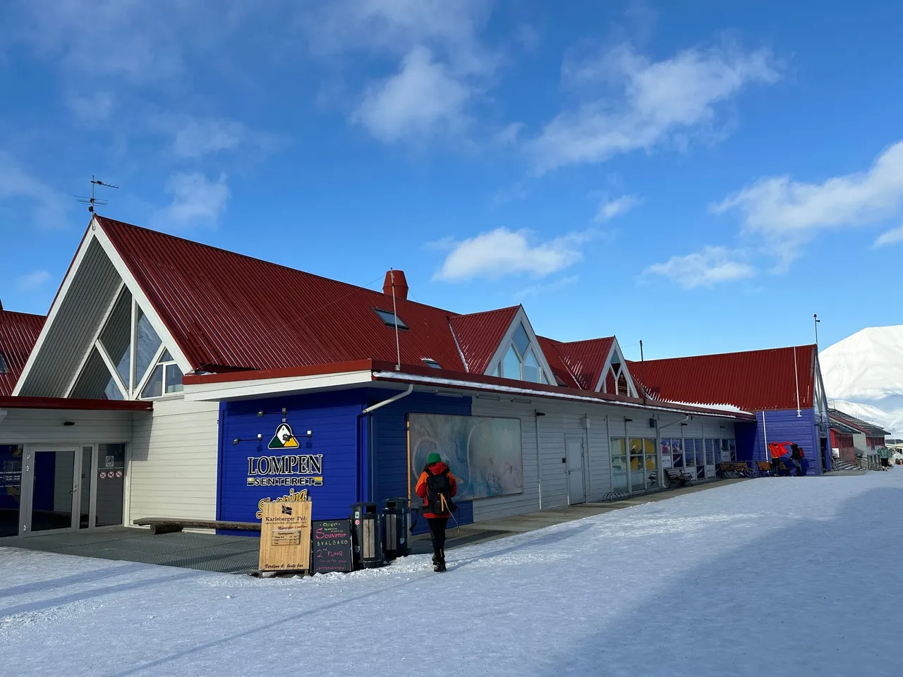 The Lompen shopping centre in Longyearbyen, a long low building with red roofs and a blue entrance, on a sunny winter day