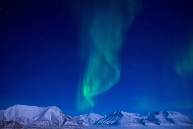 Green northern lights curling across a deep blue night sky above a row of snow-covered mountains